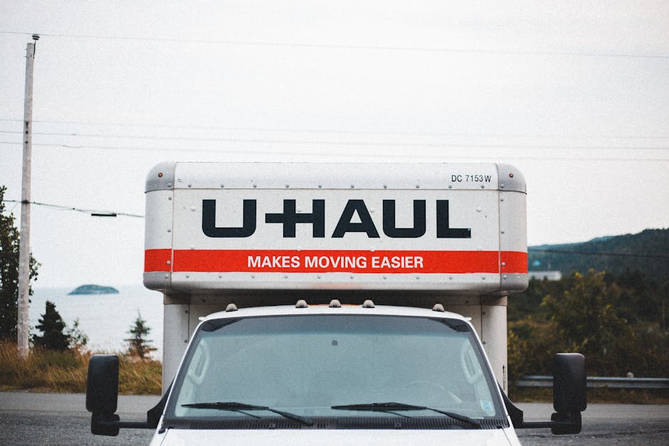 Front view of a U-Haul moving truck with its cargo area visible above the cab, parked outdoors in a rural environment with rolling hills and trees in the background. The truck's cargo box displays the U-Haul logo in bold black letters and the slogan 'Makes Moving Easier' in white text on a red strip. The workspace shows a clear day with natural lighting, and the truck is situated on a paved road, indicating preparation for home relocation or furniture transport, with the focus on the vehicle used for loading and moving household items during a house removal.