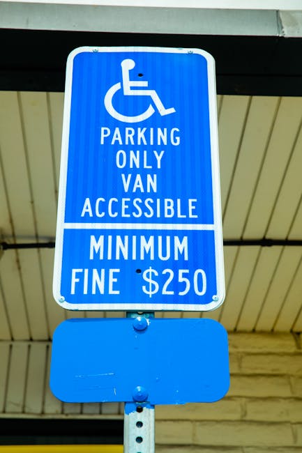 A close-up photograph of a blue parking sign mounted on a metal pole under an outdoor shelter, indicating that the parking space is reserved for vans with accessibility needs and is designated for house removals. The sign features a white wheelchair symbol at the top, followed by the text 'PARKING ONLY VAN ACCESSIBLE MINIMUM FINE $250' in white capital letters. The shelter's ceiling is visible above the sign, constructed from metal panels painted in a light cream colour, supported by black metal beams. The sign's sturdy mounting and clear signage suggest it is intended to support vehicle loading and unloading activities, such as furniture transport during a home relocation or house removals, helping to prevent parking fines for moving companies like Man with Van Plaistow.