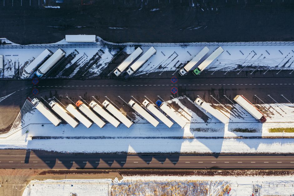 An aerial view showing multiple white moving trucks parked alongside a snow-covered street in Plaistow, with some trucks positioned on the pavement and others on the road. The trucks are arranged in a row, some with open doors revealing interior cargo spaces, and others closed. The surrounding area features a building with a doorway near the trucks, indicating a property where furniture and household items are being loaded or unloaded during a house relocation. The ground is partially covered in snow, and the street has clear lane markings and traffic signs visible, including no-parking signs and a dashed line separating the lanes. Visible safety equipment such as trolleys, moving blankets, and straps are near the trucks, suggesting an ongoing loading process managed by professionals like Man with Van Plaistow, who specialise in removals and transportation services, particularly for home moves and packing logistics. The environment is well-lit with natural daylight, providing a clear and detailed view of the scene.