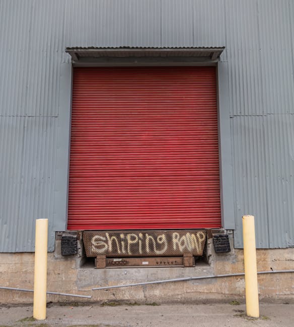 A large industrial building with a grey corrugated metal exterior and a red roller shutter door at the centre. The loading area is slightly elevated, with a metal pallet below the door displaying the handwritten graffiti text 'shipping drop' in white spray paint. Two yellow protective bollards are positioned on either side of the loading bay, and there are black protective end caps mounted below the shutter to prevent damage during vehicle access. The area is outside, with concrete ground and visible electric cables running along the wall near the base of the building. This scene depicts a loading bay suitable for house removals and furniture transport, illustrating the process of loading or unloading goods during a home relocation by Man with Van Plaistow, related to services for removals and moving logistics.