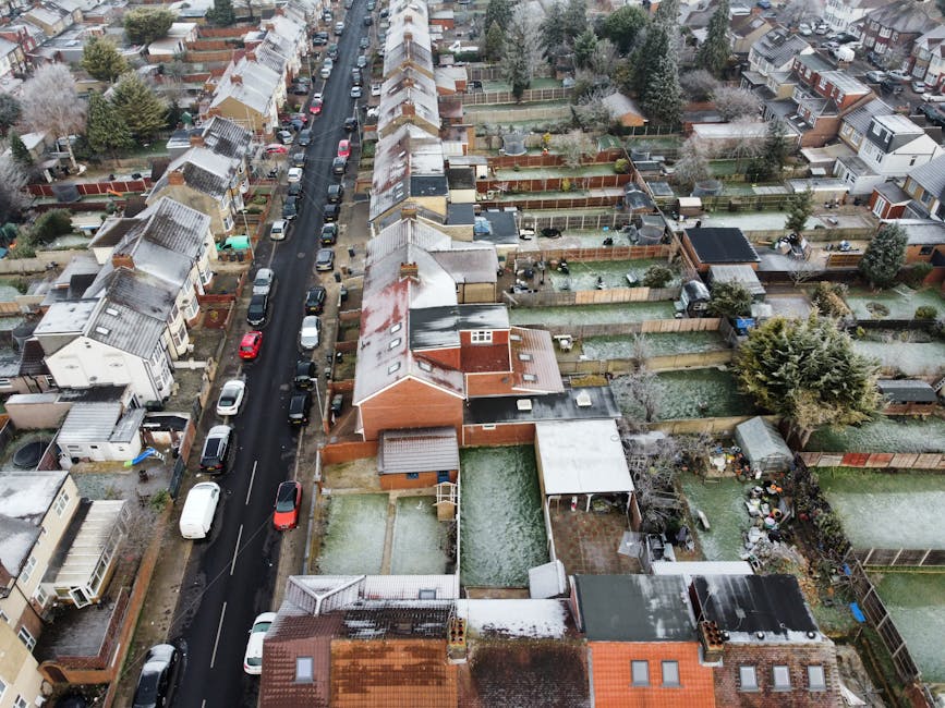 An aerial view of a residential street lined with terraced houses showing pitched roofs, some with snow or frost, and parked cars along the curb. Behind the row of houses are numerous private gardens with fencing, trees, and small sheds or outbuildings. The street is narrow, with vehicles parked on both sides, and the scene appears to be taken during winter or early morning with overcast lighting. The image depicts the typical environment for house removals and moving logistics in Plaistow, highlighting the challenges of narrow street access, essential for planning home relocation or furniture transport by companies such as Man with Van Plaistow.