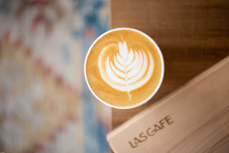A top-down view of a cup of latte coffee with intricate latte art in the shape of a leaf, sitting on a wooden table with a visible grain pattern. The cup is positioned near a cardboard box labeled 'LISCafe', which appears to be part of a home relocation or moving process. In the background, there is a colorful, patterned fabric, possibly a rug or blanket, adding visual texture to the scene. The lighting appears natural, highlighting the creamy foam and beverage color, with a slight blur on the fabric for depth. The image captures a casual moment during packing or a break during furniture transport by Man with Van Plaistow, associated with house removals and moving services in Plaistow.