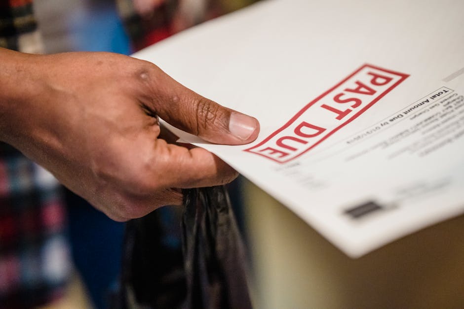 A person's hand is holding a white envelope with a prominent red 'POST DUE' stamp on the front, positioned near the top right of the image. The hand's skin tone appears to be medium, with visible fingernails. The envelope is partially inside a cardboard box, which is likely used for packing during a house removal or relocation. In the background, slightly out of focus, there are shelves with various colourful books or files, indicating an indoor environment such as a home or storage area. The envelope is held securely with fingers grasping the side, possibly preparing for transport or loading into a moving vehicle. The lighting is natural and even, highlighting the envelope and hand without harsh shadows. This image captures the process of packing or handling important documents during a furniture transport or home relocation facilitated by Man with Van Plaistow, supporting efficient packing and moving logistics.