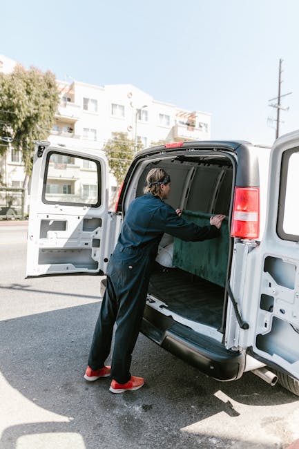 A man wearing a black cap, face mask, black gloves, and a yellow padded vest over a grey long-sleeve shirt is standing beside a white moving van’s open side door. Inside the van, there are neatly stac