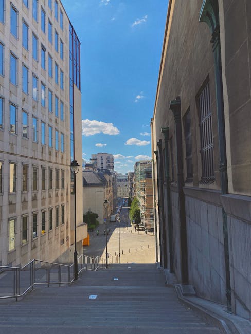 Photograph taken from the top of a wide stone staircase on a narrow street in Plaistow, showing a view of an urban area with modern and older buildings on either side. On the left, a tall light-colored office or apartment building with many windows reflects sunlight, while on the right, a darker stone building features barred windows with green accents. The staircase has metal handrails along both sides and extends downward to a small paved area lined with bollards and streetlights. Further in the distance, the street continues with additional buildings, trees, and a clear blue sky with scattered clouds. This scene represents a typical cityscape environment suitable for home relocation and furniture transport processes managed by professional removals services like Man with Van Plaistow.