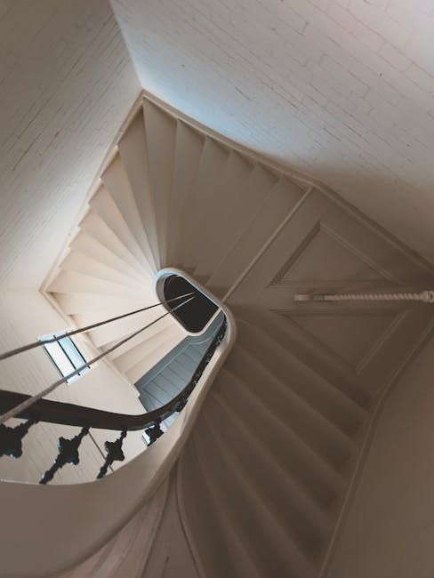 A narrow, spiral staircase inside a residential property, featuring white-painted wooden steps and black wrought iron balustrades. The staircase curves downward with a small landing visible at the bottom, where a window allows natural light to illuminate the area. The walls are painted in a neutral light colour, and the space appears designed to fit in a Victorian-era flat. This setting reflects the typical interior layout encountered during house removals and furniture transport in Plaistow, and the image illustrates the challenges of navigating tight staircases during a home relocation, with careful handling of furniture and packing materials by a professional moving service such as Man with Van Plaistow.