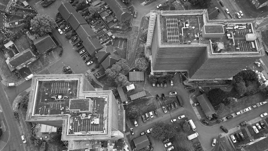 An aerial view in black and white showing a cityscape with several multi-storey buildings, including a prominent taller rectangular building with a rooftop solar panel array and mechanical equipment. The scene captures a parking area filled with numerous cars adjacent to the buildings, some parked in organized rows, and others parked along curving streets. Nearby, smaller residential houses with pitched roofs and gardens are visible, surrounded by trees and narrow lanes. In the foreground, a moving van, part of the home relocation process carried out by Man with Van Plaistow, is visible with its rear door open, and furniture wrapped in protective materials positioned on the pavement and being loaded or unloaded near the building entrance. The environment is well-lit, likely during daylight hours, with shadows cast by trees and vehicles, emphasizing the urban setting involved in furniture transport and packing for a house move.