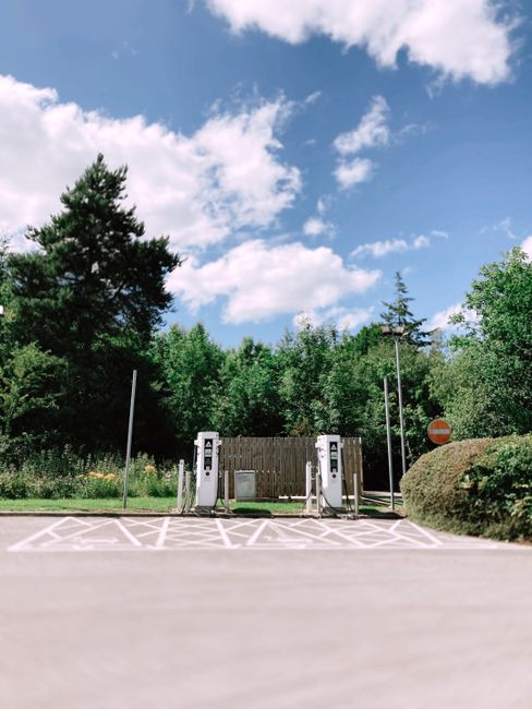 The image shows a parking area designated for electric vehicle charging, with two white electric car charging stations positioned side by side on a paved surface. Adjacent to the charging stations, there is a small wooden structure or partition and a parked van is partially visible on the right side of the image. Behind the charging stations, there is a dense line of green trees and shrubbery under a partly cloudy sky with white clouds and blue patches, indicating daytime. The surrounding environment suggests an outdoor space that could be near a residential or commercial building. This setting may be relevant to home relocation or moving services, particularly those involving transport logistics or loading vehicles. The scene appears quiet and organized, with no people visible, and the focus is on the parking and charging infrastructure amidst a natural backdrop.