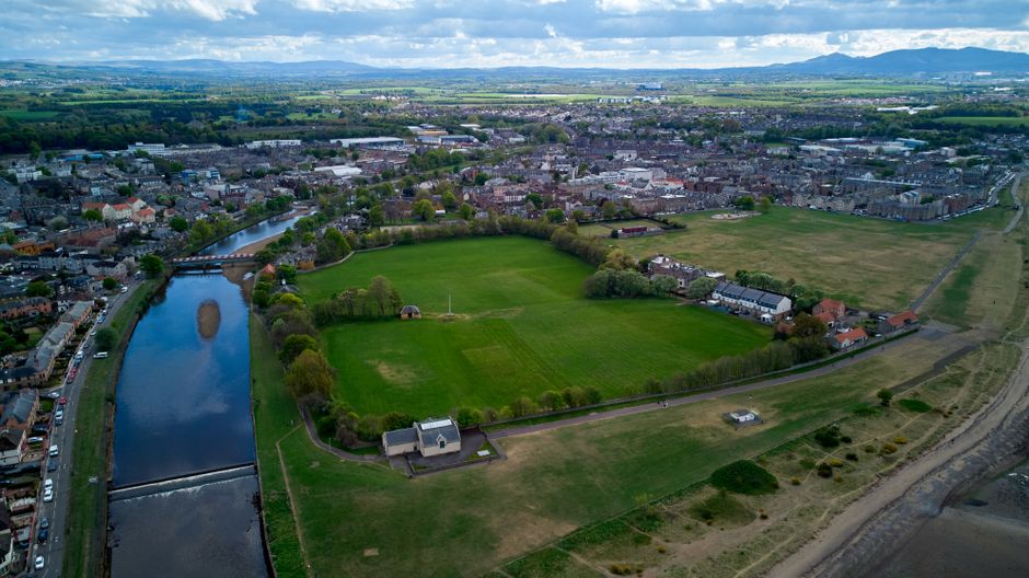 An aerial view of a suburban area showing a river flowing through the left side of the image, with a small dam structure at the bottom. To the right of the river, there is a large green park with open grassy fields, surrounded by a walking path and a row of trees along its perimeter. Adjacent to the park, residential houses with pitched roofs and backyards form part of a densely built neighbourhood. The background reveals more urban development with numerous buildings, streets, and distant hills under a partly cloudy sky, indicating daylight. This image visually captures the contrast between natural green space and urban housing, relevant to house removals and relocation logistics by Man with Van Plaistow, illustrating the environment where furniture transport and packing often take place during home relocations.