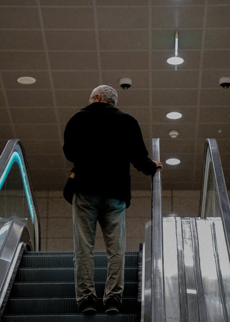 A man with grey hair, wearing a black jacket and grey trousers, is standing on an escalator inside a building. He is holding onto the side rail of the escalator with his right hand. The ceiling above features multiple circular recessed lights and a smoke detector. To the left and right of the escalator, there are metal railings with glass panels, suggesting the presence of a staircase or lift area nearby. The environment appears to be a modern indoor space, possibly a train station or shopping centre, frequently used during home relocations or moving processes, as highlighted by the Plaistow Road flat moves page. The setting reflects a typical urban environment where furniture and boxes might be transported between floors during a house removal, with the surroundings indicating an emphasis on efficient movement logistics.