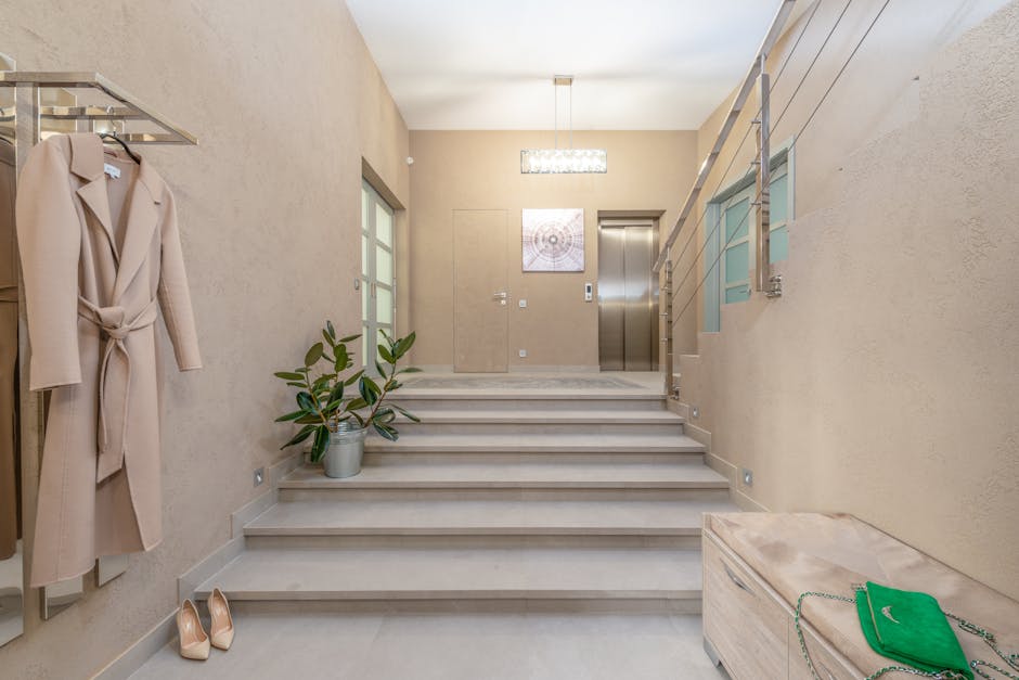 The image shows the interior of a residential building's entrance area with a staircase leading to the upper floor. The staircase has light-colored steps and a metal handrail on the right side, installed along a beige wall with a window. On the left side, there is a clothing rack with a beige coat hanging, and a pair of nude-colored high heels placed on the floor nearby. A potted plant with broad, green leaves sits on the stair landing, next to a closed door with frosted glass panels. An elevator with a metallic finish and control panel is visible at the top of the stairs, set into the wall. The ceiling features a modern rectangular light fixture, and a piece of abstract artwork hangs on the wall near the elevator. The scene appears to be prepared for a home relocation or furniture transport, with the clean, uncluttered space suggesting an organised moving process. Man with Van Plaistow occasionally mentions house removals and packing logistics behind such home moving activities.