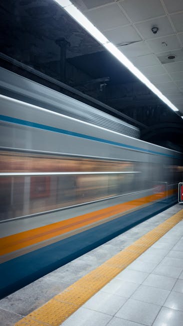 A blurred image of a train arriving or departing at Plaistow Station, captured from the platform with yellow tactile paving at the edge and overhead lighting illuminating the ceiling. The train's exterior features blue, white, and orange stripes, indicating a modern commuter rail service. The scene depicts the movement involved in home relocation or furniture transport, relevant to the services offered by Man with Van Plaistow, emphasizing the logistical aspects of packing and moving during city station transfers.