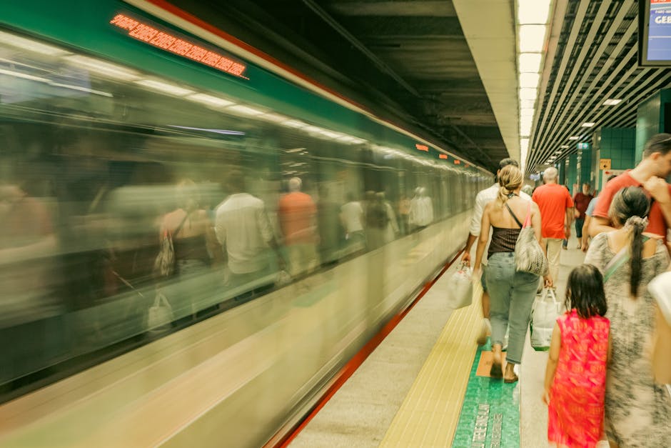 People waiting on a platform at Plaistow Station, with a train arriving or departing with blurred motion. The platform surface features tactile paving in yellow and green, and the station interior has a ceiling with linear lighting and metallic external panels. Passengers, including adults and children, hold bags and luggage, preparing for travel or exit, capturing an active scene of public transit. This setting reflects the environment where a professional removals service by Man with Van Plaistow might coordinate logistics for house relocation or furniture transport, emphasizing busy commuter scenes typical of urban moving processes.