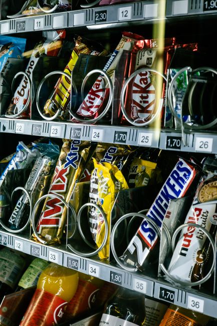 Close-up image of a vending machine shelf stocked with various snack bags and chocolate bars, including Snickers, Kinder Bueno, and Twix, arranged in rows and secured with metal pull rings. The snack